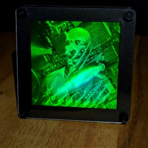 May include: A black, square-framed display shows a green holographic image of a human face and circuit board patterns. The image is illuminated with a bright green light, creating a futuristic and technological aesthetic. The display is mounted on a wooden surface.