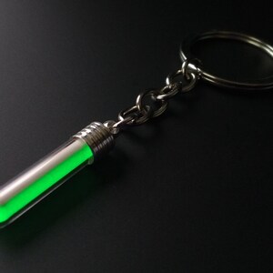 Puede incluir: Un llavero con un peque&ntilde;o tubo de vidrio transparente que contiene una sustancia verde brillante. El tubo est&aacute; conectado a una cadena y un anillo de color plateado. El objeto est&aacute; sobre un fondo oscuro.