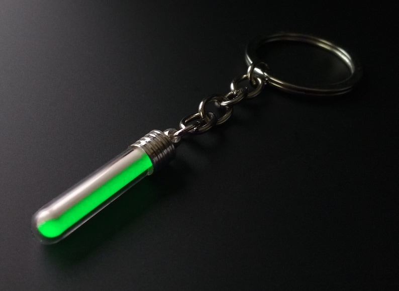 Puede incluir: Un llavero con un peque&ntilde;o tubo de vidrio transparente que contiene una sustancia verde brillante. El tubo est&aacute; conectado a una cadena y un anillo de color plateado. El objeto est&aacute; sobre un fondo oscuro.