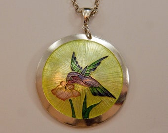 May include: A silver necklace with a round pendant featuring a green, purple, and pink hummingbird with a yellow flower. The pendant has a textured, sunburst design.