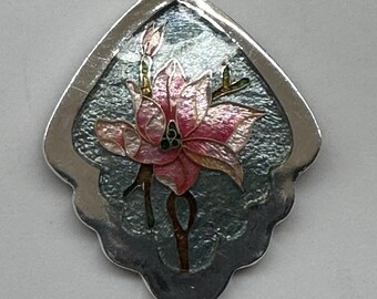 May include: A silver pendant necklace with a delicate, scalloped, leaf-shaped design. The pendant features a pink flower with green leaves and a silver stem, all set against a blue and white enamel background.