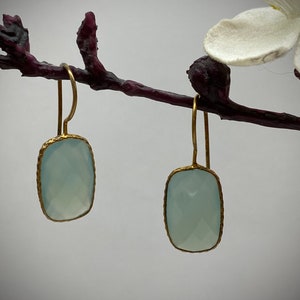 Puede incluir: Un par de pendientes de oro con gotas de piedras preciosas de color azul claro. Los pendientes están colgados de una rama.