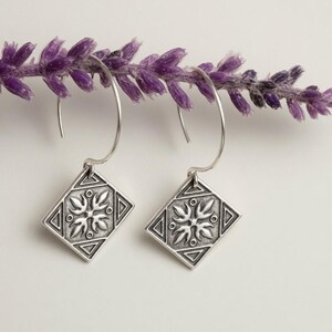 May include: A pair of silver drop earrings featuring a diamond-shaped pendant with an embossed floral motif and geometric border. The earrings hang from simple silver hooks, showcasing a polished finish with darkened recessed details.