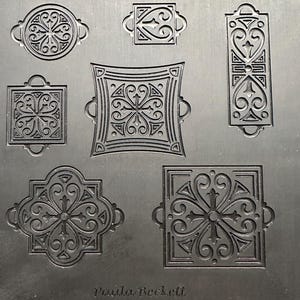 Op de afbeelding: Een metalen plaat met zeven in reliëf gemaakte decoratieve ontwerpen. De ontwerpen omvatten cirkels, vierkanten en rechthoeken, elk met ingewikkelde bloemen- en krulpatronen. De naam "Paula Beckett" staat onderaan afgedrukt.