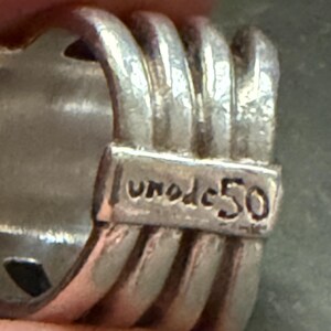 Pode incluir: Close-up de um anel de prata com um design texturizado. O anel apresenta uma s&eacute;rie de cristas paralelas e o texto gravado "uno de 50". O anel &eacute; feito de um material met&aacute;lico brilhante.