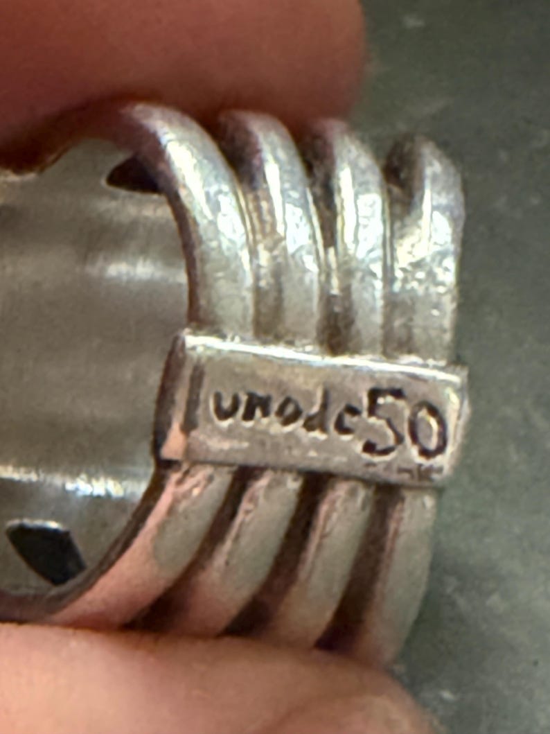 Pode incluir: Close-up de um anel de prata com um design texturizado. O anel apresenta uma s&eacute;rie de cristas paralelas e o texto gravado "uno de 50". O anel &eacute; feito de um material met&aacute;lico brilhante.