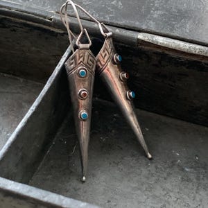 May include: A pair of silver-toned earrings with a conical design. Each earring features a geometric pattern at the top and is adorned with small, colorful gemstones in turquoise and red. The earrings have a hook closure.