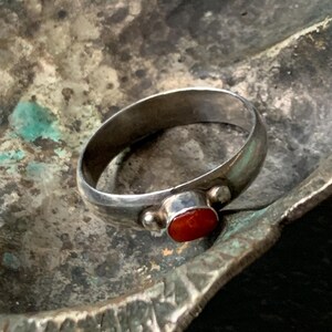 May include: A silver ring with a small, oval, orange-red stone. The ring has a simple band and two small silver beads on either side of the stone. The ring is displayed on a textured, metallic surface.