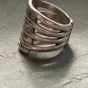 Pode incluir: Um anel prateado com um design &uacute;nico. O anel apresenta m&uacute;ltiplas faixas horizontais conectadas por barras verticais, criando uma estrutura aberta em forma de gaiola. O anel &eacute; feito de metal brilhante.