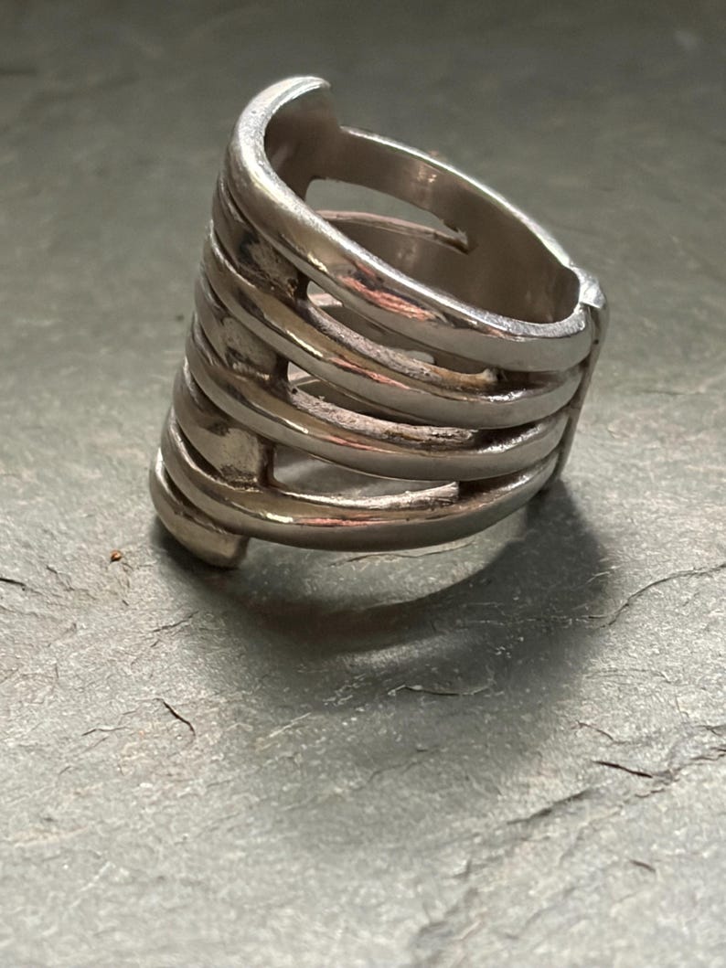 Pode incluir: Um anel prateado com um design &uacute;nico. O anel apresenta m&uacute;ltiplas faixas horizontais conectadas por barras verticais, criando uma estrutura aberta em forma de gaiola. O anel &eacute; feito de metal brilhante.