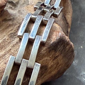 Puede incluir: Una pulsera de tono plateado que presenta una serie de eslabones rectangulares de metal pulido conectados en un patrón repetitivo. Los eslabones están dispuestos en un diseño lineal y estructurado con una superficie lisa y reflectante.