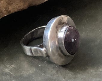 May include: A silver ring with a large, round, dark purple gemstone set in a bezel. The ring has a wide band with a textured design near the base. The gemstone is the focal point, set in a circular, raised setting.