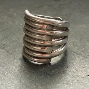 Pode incluir: Um anel prateado com um design &uacute;nico. O anel apresenta v&aacute;rias faixas curvas empilhadas, criando uma apar&ecirc;ncia texturizada e atraente. As faixas s&atilde;o conectadas por pequenos elementos quadrados. O anel &eacute; uma pe&ccedil;a de joalharia marcante.