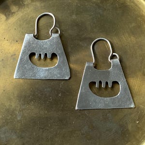 May include: A pair of silver-toned earrings. Each earring has a rectangular shape with a curved top and a cutout design near the bottom. The earrings are displayed on a gold-colored surface.