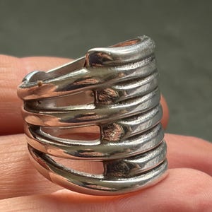 Pode incluir: Um anel prateado com um design &uacute;nico. O anel apresenta m&uacute;ltiplas faixas horizontais conectadas por barras verticais, criando uma estrutura aberta em forma de gaiola. O metal tem um acabamento polido, refletindo a luz.