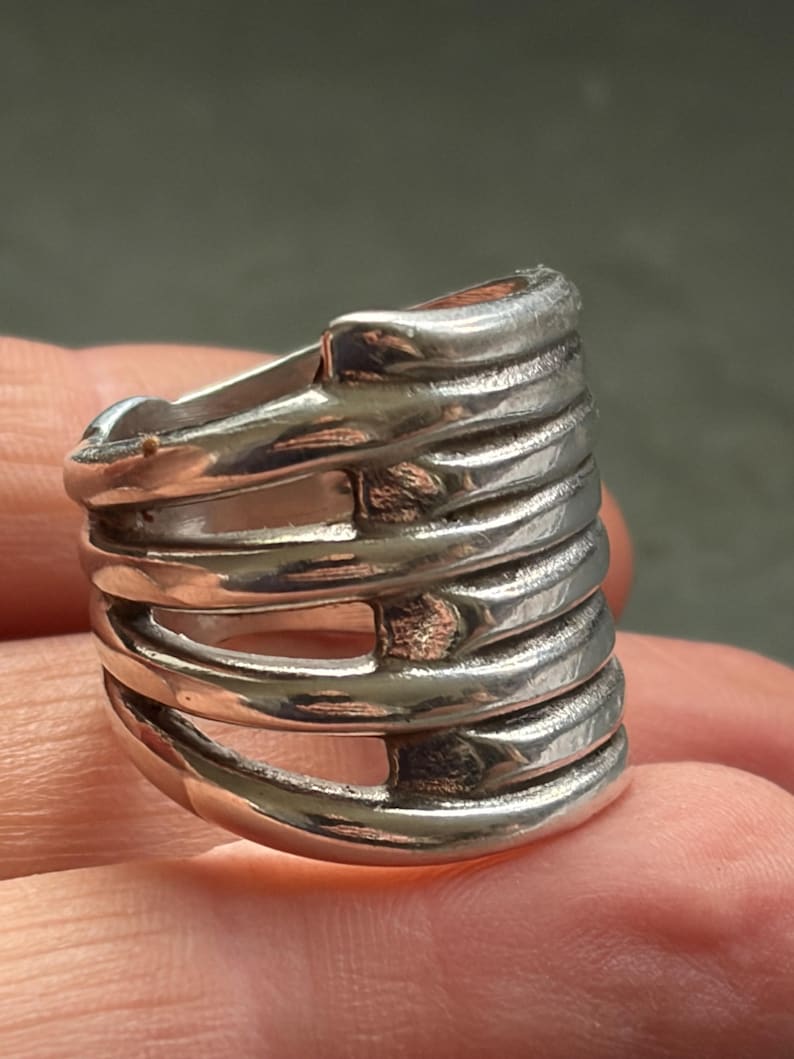 Pode incluir: Um anel prateado com um design &uacute;nico. O anel apresenta m&uacute;ltiplas faixas horizontais conectadas por barras verticais, criando uma estrutura aberta em forma de gaiola. O metal tem um acabamento polido, refletindo a luz.