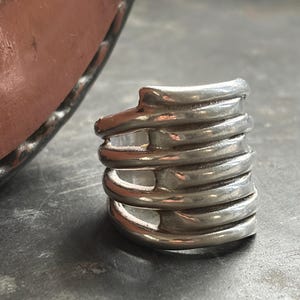 Pode incluir: Um anel prateado com um design &uacute;nico e em camadas. O anel apresenta v&aacute;rias faixas curvas empilhadas umas sobre as outras, criando uma apar&ecirc;ncia texturizada e dimensional. O metal tem um acabamento polido, refletindo a luz e aumentando seu apelo visual.