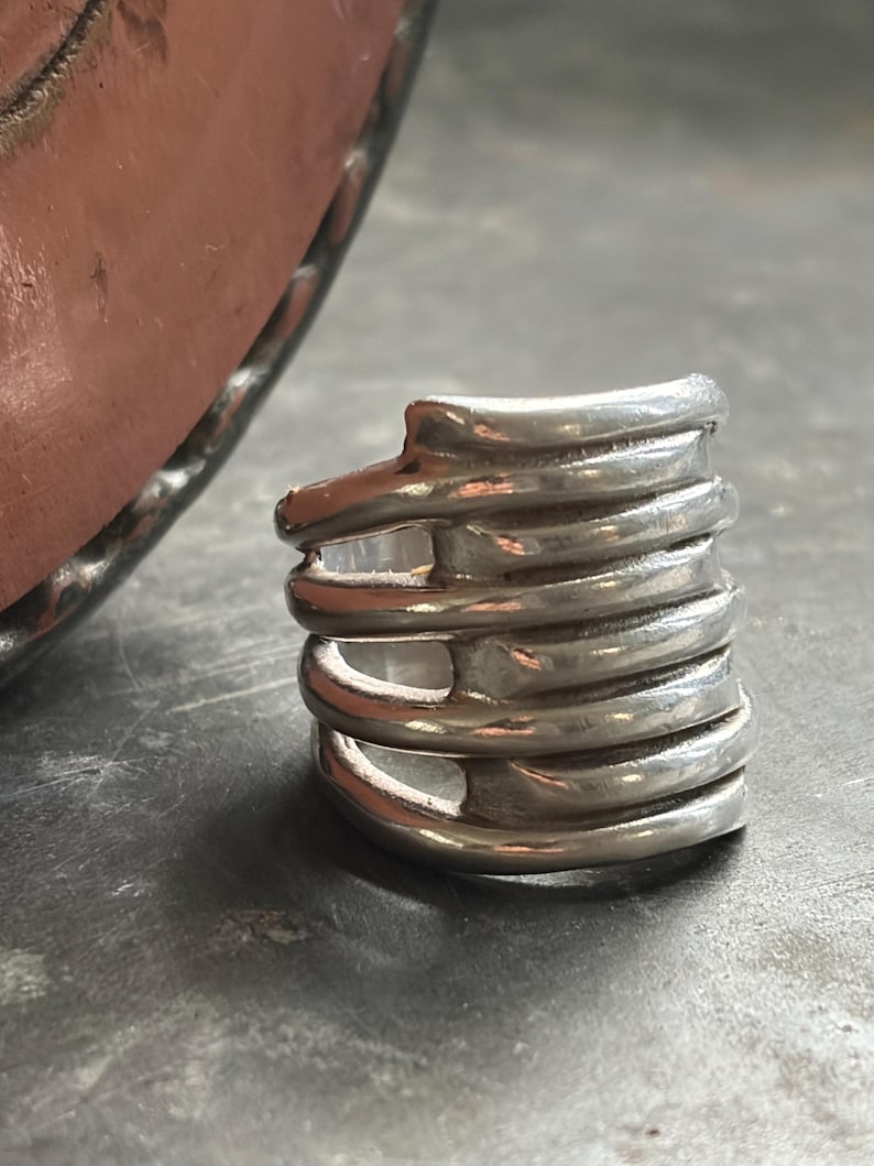 Pode incluir: Um anel prateado com um design &uacute;nico e em camadas. O anel apresenta v&aacute;rias faixas curvas empilhadas umas sobre as outras, criando uma apar&ecirc;ncia texturizada e dimensional. O metal tem um acabamento polido, refletindo a luz e aumentando seu apelo visual.