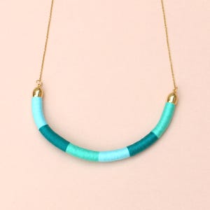 Puede incluir: Un collar colorido con un colgante curvo y cilíndrico. El colgante está envuelto en hilos de color turquesa, turquesa y azul claro. El collar tiene una delicada cadena dorada y extremos dorados.