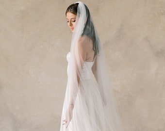 May include: A bride wearing a white strapless wedding dress with a long, flowing veil. The veil is made of tulle and is very sheer. The bride is standing in front of a beige background.