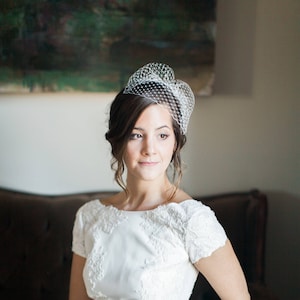 May include: A bride wearing a white lace wedding dress with short sleeves and a birdcage veil. She is sitting on a brown sofa and looking to the right.