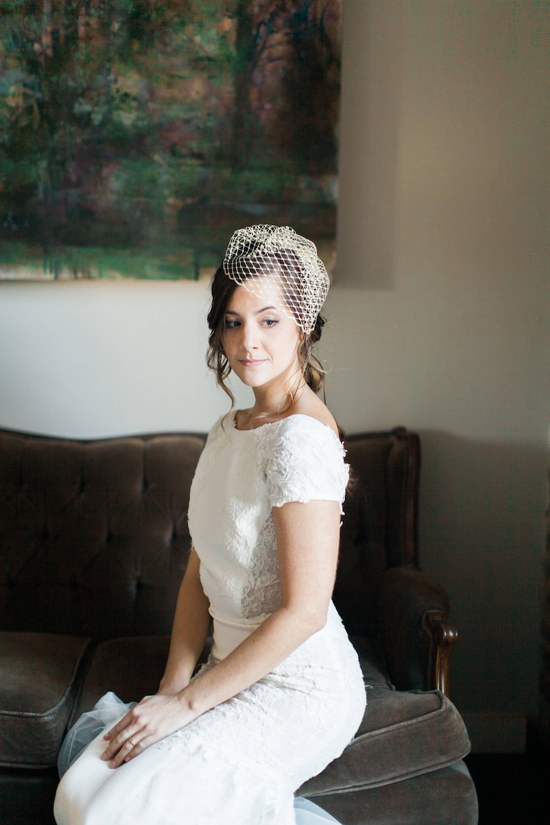 May include: A bride wearing a white lace wedding dress with short sleeves and a veil. She is sitting on a brown sofa in front of a painting.