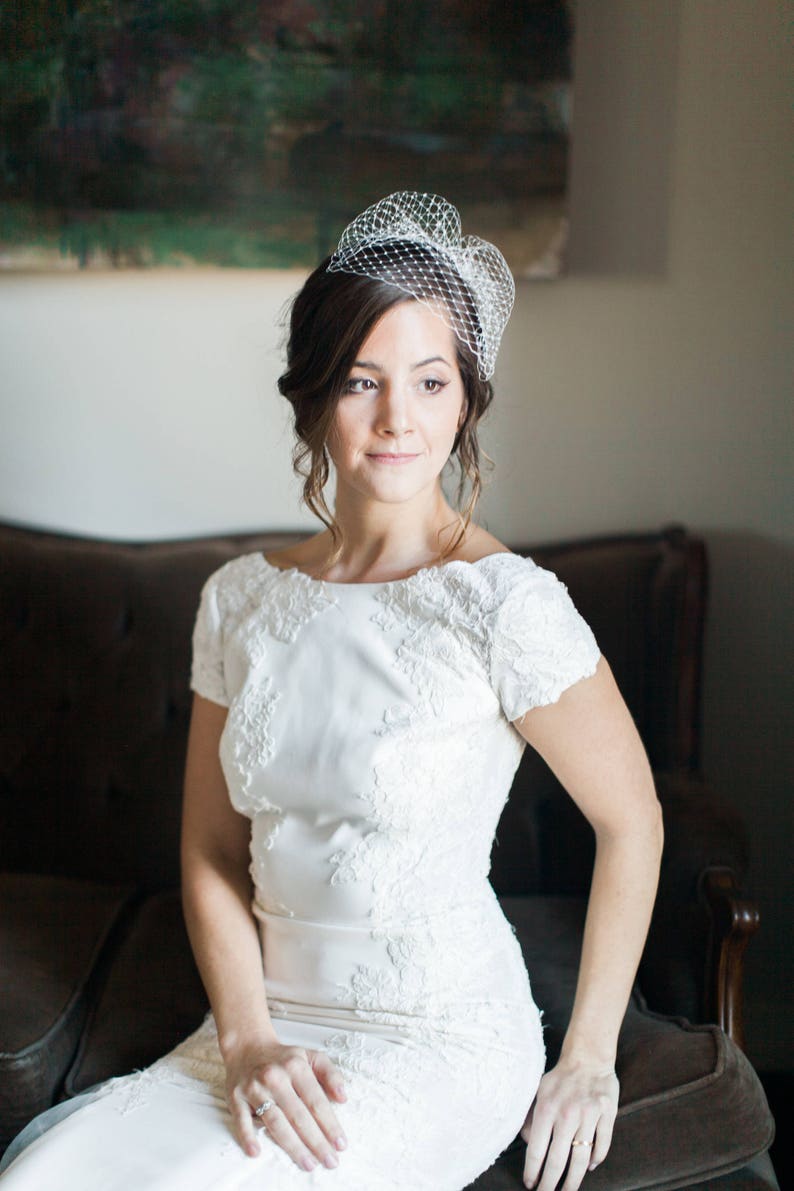 May include: A bride wearing a white lace wedding dress with short sleeves and a birdcage veil. She is sitting on a brown sofa and looking to the right.