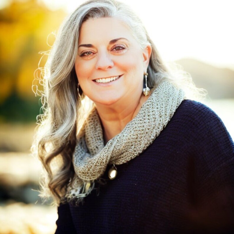 May include: A woman with long, grey hair smiles at the camera. She is wearing a dark blue jumper and a light brown scarf. The background is blurred and out of focus.