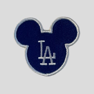 May include: A blue embroidered patch shaped like Mickey Mouse ears with the letters "LA" in white inside.
