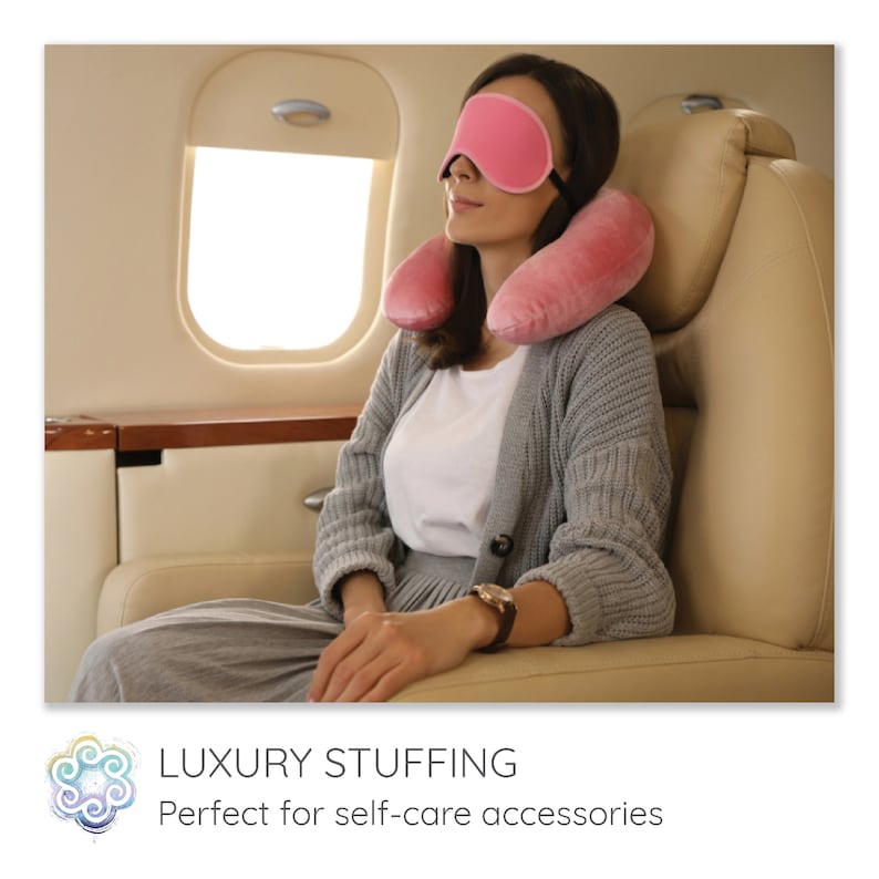 May include: A woman wearing a pink eye mask and a pink neck pillow while sitting in an airplane seat.  The seat is beige leather.  The woman is wearing a gray sweater and a watch.