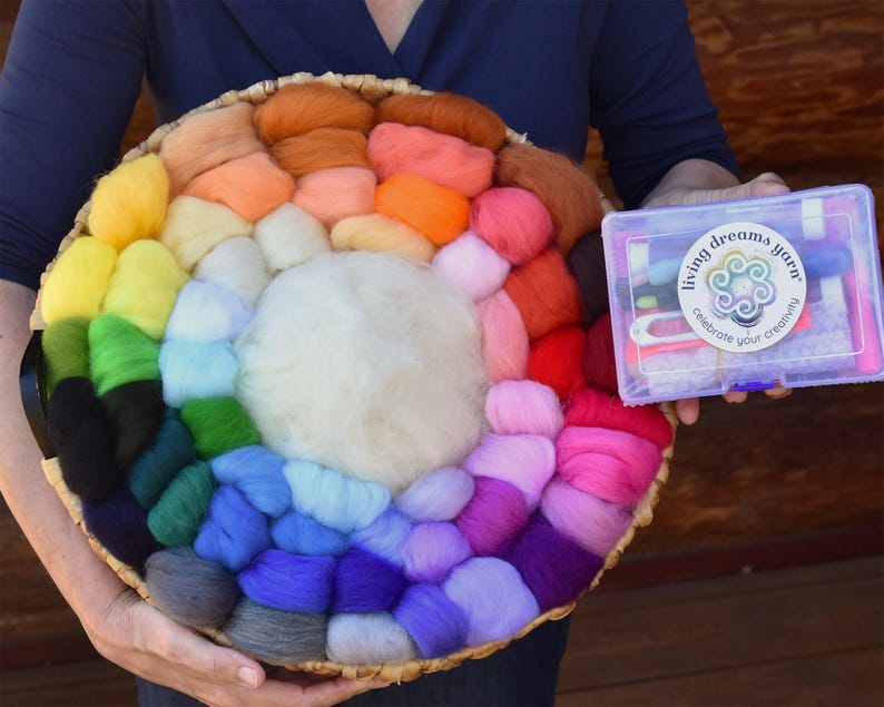 May include: A colorful assortment of wool roving arranged in a circular basket, showcasing a spectrum of hues from yellow to purple. A clear plastic box with the text "living dreams yarn" is held nearby, suggesting a crafting kit.