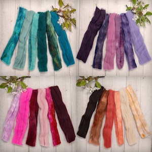 May include: Assortment of colorful wool roving fibers in various shades of blue, purple, pink, red, brown, and beige. The fibers are arranged in rows on a white wooden surface.