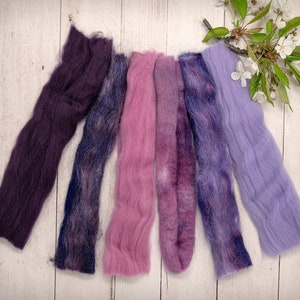 May include: Six strands of hand-dyed wool yarn in shades of purple, pink, and lavender. The yarn is arranged in a row on a white wooden surface.