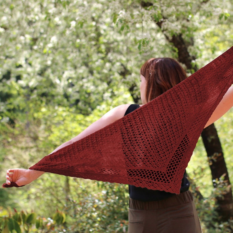 May include: A burgundy-colored, open-weave triangular shawl is held up against a blurred background of green foliage and white blossoms. The shawl's texture is visible, with a delicate, lacy pattern. The person holding the shawl is wearing a black top and brown pants.