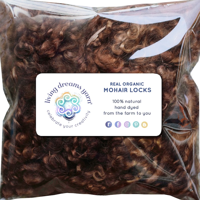 Op de afbeelding: Een doorzichtige plastic zak gevuld met bruine mohair lokken. De zak heeft een label met de tekst "Living Dreams Yarn" en "Real Organic Mohair Locks". Het label vermeldt ook dat het mohair 100% natuurlijk is, handgeverfd en rechtstreeks van de boerderij komt.