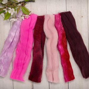 May include: Six strands of wool yarn in various shades of pink, red, and purple. The yarn is arranged in a row on a white wooden surface.