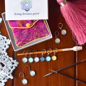 Può includere: Un set di marcatori di punto colorati con un design a spirale, una pochette rosa e due ferri da maglia. I marcatori di punto sono attaccati a un bastoncino di legno. La pochette ha il testo "living dreams yarn" sulla scatola.