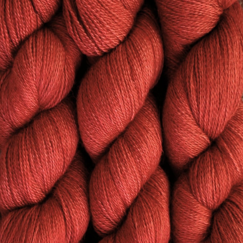 May include: Close-up of a skein of red-orange yarn. The yarn is tightly twisted, revealing a textured surface. The color is a warm, earthy tone, ideal for knitting or crocheting projects. The yarn appears soft and luxurious.