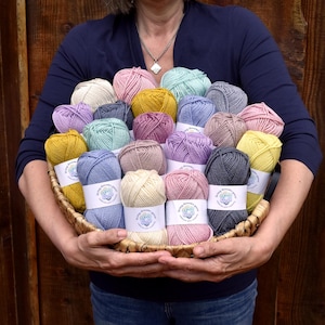 May include: A wicker basket filled with colorful yarn skeins in various pastel shades, including pink, yellow, blue, and purple. Each skein is wrapped with a white label. The basket is held by a person against a wooden background.