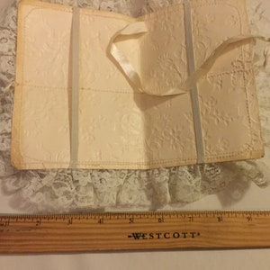 K&ouml;nnte beinhalten: Ein wei&szlig;es, mit Spitze besetztes B&uuml;chlein mit einem Satinbandverschluss. Das B&uuml;chlein ist aus dickem Papier mit einem gepr&auml;gten Blumenmuster.