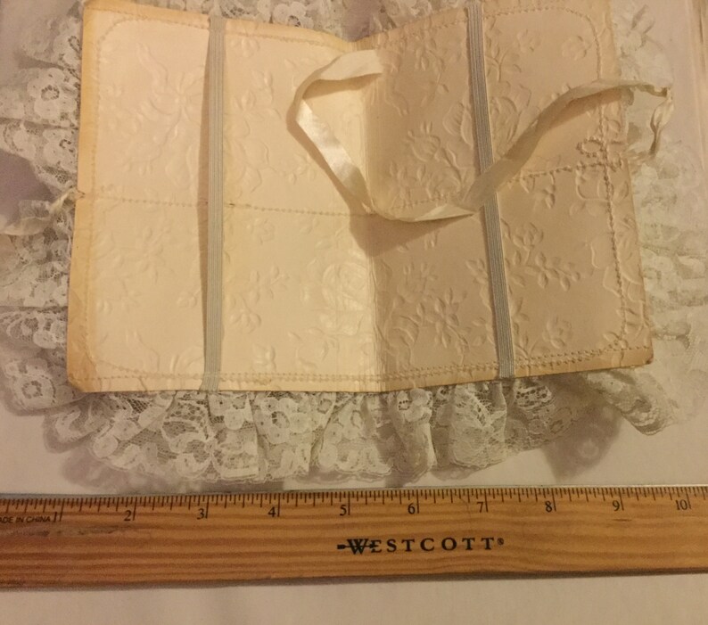 K&ouml;nnte beinhalten: Ein wei&szlig;es, mit Spitze besetztes B&uuml;chlein mit einem Satinbandverschluss. Das B&uuml;chlein ist aus dickem Papier mit einem gepr&auml;gten Blumenmuster.