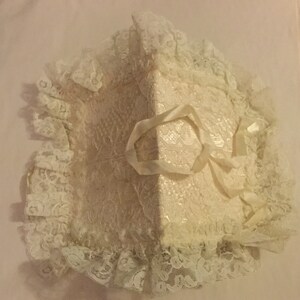 K&ouml;nnte beinhalten: Ein wei&szlig;es Kissen aus Spitze mit einem Satinband, das zu einer Schleife gebunden ist. Das Kissen ist perfekt, um Trauringe w&auml;hrend einer Zeremonie zu halten.