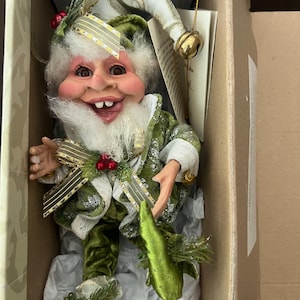 Puede incluir: Una caprichosa figura de elfo con una cara sonriente, barba blanca y ropa de terciopelo verde. El elfo lleva un gorro puntiagudo con detalles dorados y sostiene una campana dorada. Bayas rojas decorativas y cinta dorada adornan el atuendo, creando un tema festivo.