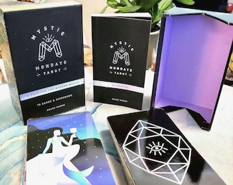May include: A black box with the text "Mystic Mondays Tarot" and a deck of tarot cards with a black and white illustration of a diamond with an eye inside. The cards are stacked and the top card shows a queen of cups.