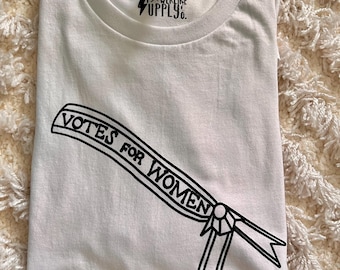 Puede incluir: Camiseta blanca con el texto "VOTES FOR WOMEN" en escritura negra sobre un diseño de pancarta. La camiseta también presenta la marca "EVERLIKE SUPPLY CO." en negro. La camiseta está doblada sobre una superficie crema texturizada.