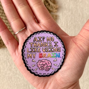 May include: A round sticker with a scalloped black border and the text "AI? NO THANKS, I LIKE USING MY BRAIN." The sticker has a light purple background with a pink brain graphic and colorful text.