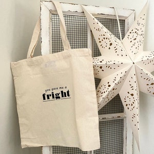 Op de afbeelding: Een witte canvas tote bag met zwarte tekst die "you gave me a fright" zegt. De tas hangt aan een witte houten deur met een witte stervormige decoratie.