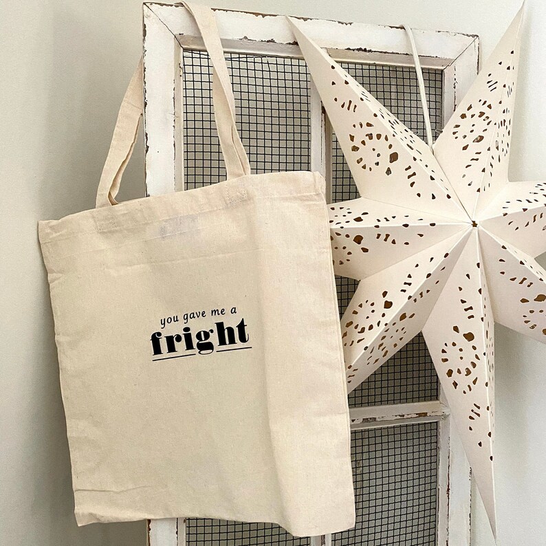 Op de afbeelding: Een witte canvas tote bag met zwarte tekst die "you gave me a fright" zegt. De tas hangt aan een witte houten deur met een witte stervormige decoratie.