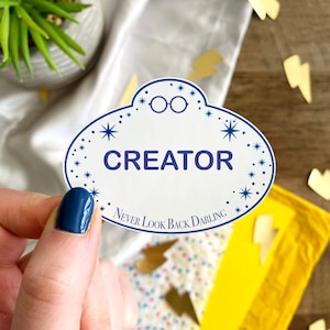 May include: White name tag with blue text and stars. The text reads "CREATOR NEVER LOOK BACK DARLING". There are glasses drawn on the tag.