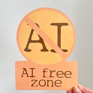 May include: A sign with a light orange background and a brown border. The sign has a circle with the letters "AI" crossed out, and the words "AI free zone" below.